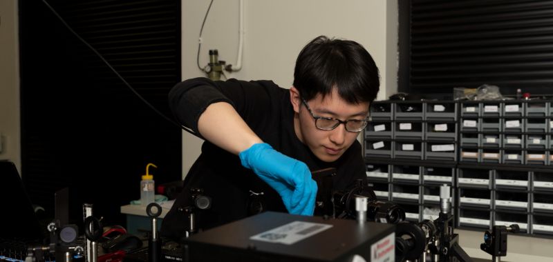 Yun Li, a member of Exciton Science based at the University of Sydney, conducts an experiment using equipment in a laser laboratory. He is wearing blue goggles, glasses and a back jumper.