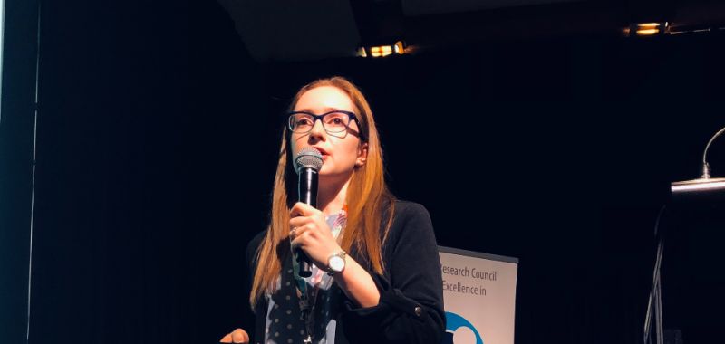 Alison Goldingay is pictured on stage holding a microphone and wearing glasses, with an Exciton Science pull-up promotional banner behind her