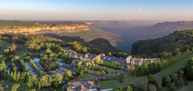 An aerial image of the Fairmont Resort and Blue Mountains, a collection of buildings above a deep, misty valley of rock faces and dense vegetation.