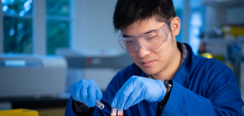 Dingchen Wen is pictured wearing a navy blue lab coat, light blue plastic gloves and clear plastic protective goggles while holding chemicals in a laboratory at the University of Melbourne
