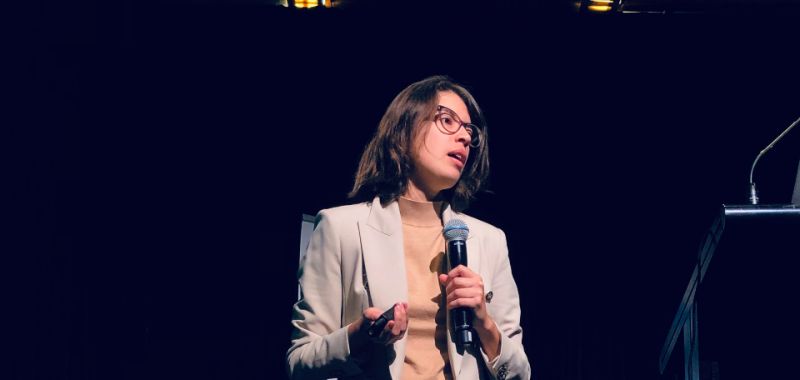 Jessica Alves standing on stage, holding a microphone. She is wearing a cream blazer, dark trousers and glasses.