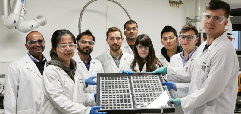 A group of Exciton Science researchers wearing white lab coats and protective goggles are pictured in a lab holding up a solar panel