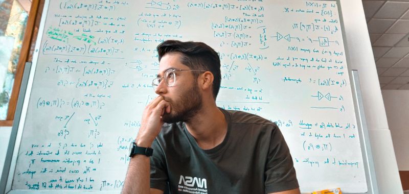 Roberto Muñoz is pictured wearing glasses and a t-shirt, in front of a large white board covered in equations written in blue.