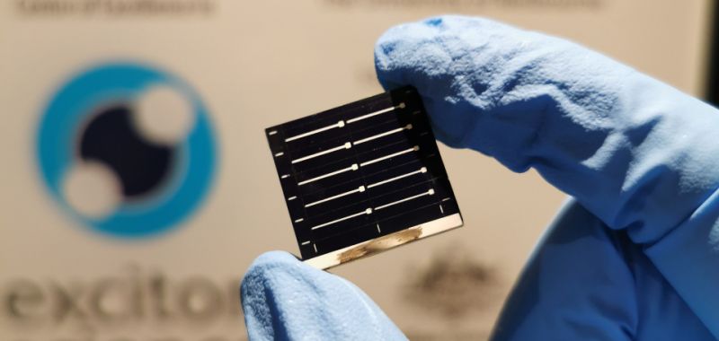 A blue-gloved hand holding a black solar cell against a white background