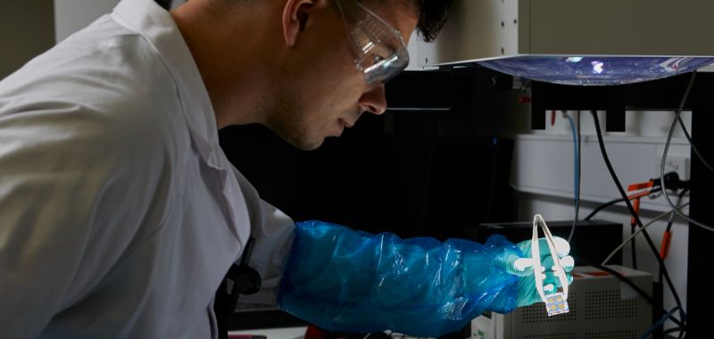 A member of Exciton Science working in a lab, wearing a white coat, blue gloves and clear plastic protective goggles