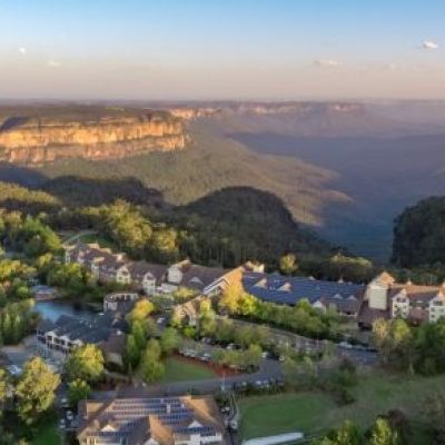 An aerial image of the Fairmont Resort and Blue Mountains, a collection of buildings above a deep, misty valley of rock faces and dense vegetation.