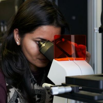 A picture of a female Exciton Science researcher using a microscope. The scientist has dark hair and is wearing red clothing.