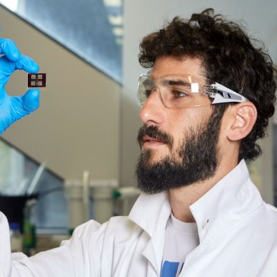 Amit Kessel of Monash University is pictured wearing a white lab coat, blue gloves, protective goggles and holding a solar cell