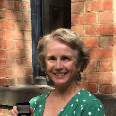 Ann Roberts is pictured wearing a green shirt and holding up a small medal in a blue display box. She is standing in front of a red brick wall outside in a courtyard filled with sunlight.