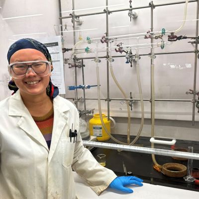 Carol Hua wearing a lab coat, goggles and blue gloves, facing the camera and smiling
