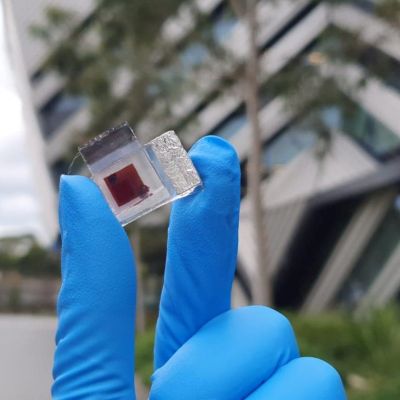 A gloved hand holds a small dye-sensitised solar cell device in front of a building at Monash University