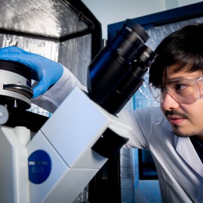 Dr Eser Akinoglu working with a microscope and wearing safety goggles and a white lab coat at The University of Melbourne