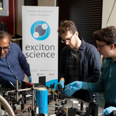 Girish Lakhwani (left) is pictured in a blue shirt working with equipment and two other colleagues in a laboratory at the University of Sydney. An Exciton Science pull-up promotional banner is in the background