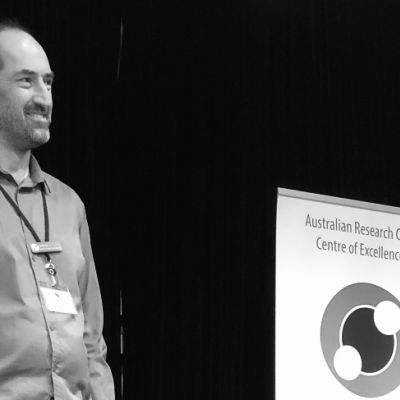Jared Cole (left) is pictured standing on stage with his hands in his pockets, smiling and focusing on something out of shot. To his right is an Exciton Science promotional banner. Jared is wearing a shirt, jeans and a lanyard. The image is in monochrome.