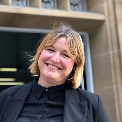 Kate McGeoch smiling and looking at the camera while wearing a black outfit and standing in front of the Chemistry building at the University of Melbourne