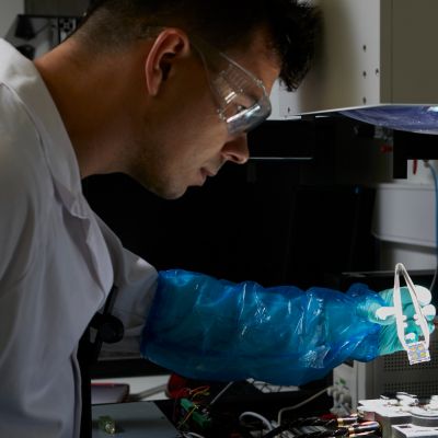 A member of Exciton Science working in a lab, wearing a white coat, blue gloves and clear plastic protective goggles