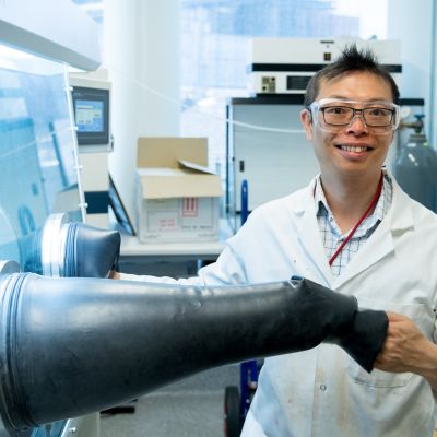 Wallace Wong is pictured wearing a white lab coat and clear plastic goggles while working with a glovebox in a laboratory