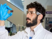 Amit Kessel of Monash University is pictured wearing a white lab coat, blue gloves, protective goggles and holding a solar cell
