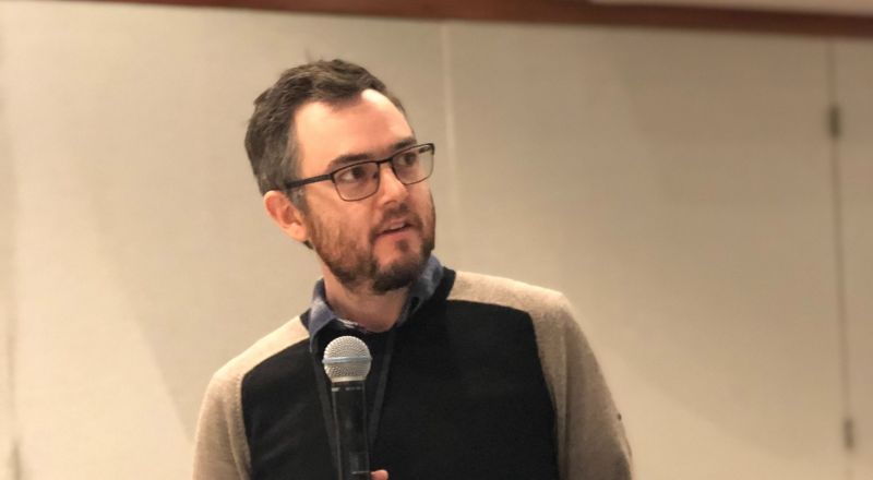Chris Hall is pictured standing and holding a microphone while looking at a projector screen out of shot. He is wearing glasses, a dark jumper and shirt. 