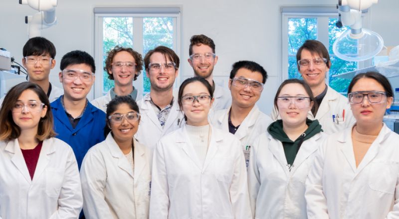 Members of Exciton Science wearing white lab coats and protective goggles stand in a group inside a laboratory and face the camera.
